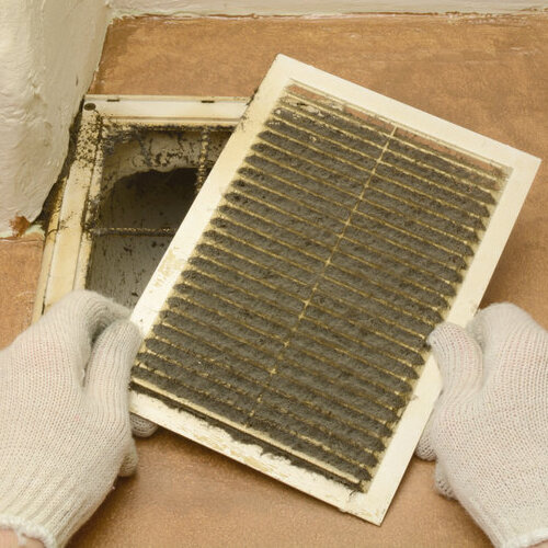 A person wearing gloves removes a dirty air vent cover as part of a heating maintenance checklist. The image emphasizes the importance of cleaning or replacing air filters and vents to maintain optimal heating system performance and indoor air quality