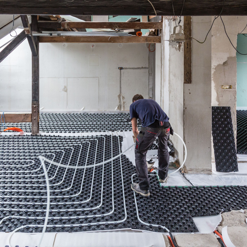 "A technician installing a radiant heating system in a large room, carefully positioning flexible heating tubes over a grid system. The image highlights the process of laying out radiant heating for efficient and even floor heating in modern construction.