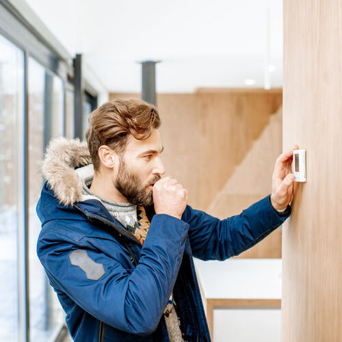 Man in winter clothes feeling cold adjusting room temperature with electronic thermostat at home