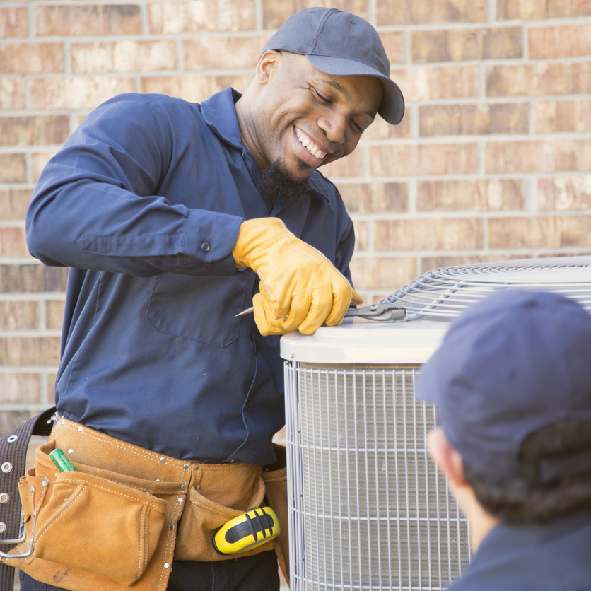 A smiling HVAC technician in a blue uniform and yellow gloves works on an air conditioning unit outside a brick building. This image highlights the Benefits of Professional AC Installation, ensuring proper setup and long-term efficiency.